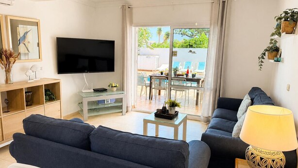A modern living room with a sofa, TV, and a dining area visible through glass doors.