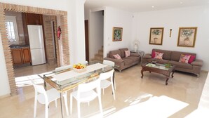 A modern living room with a glass dining table, white chairs, a brown sofa, and a coffee table. There is a kitchen area with a refrigerator and a brick wall.