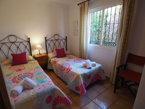 A room with two beds, each with a floral bedspread and a red pillow. There is a window with a view of greenery outside, and a chair is visible in the corner. - Nerja Paradise Rentals - Villa Los Girasoles (Nerja)
