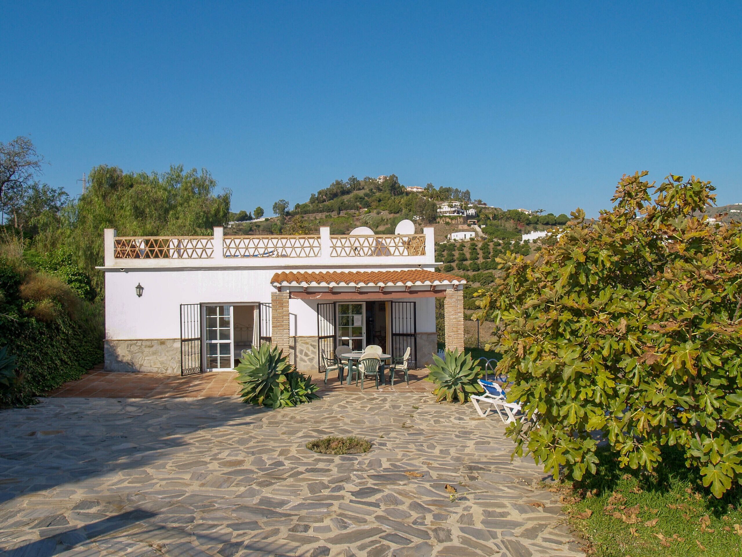 Una casa de dues plantes amb un pati de pedra, un balcó amb baranes de gelosia i vistes als turons circumdants.