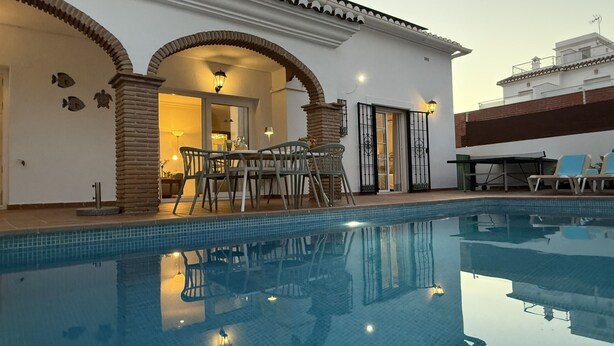 Una piscina con zona de patio, una mesa de comedor con sillas y vistas a un edificio con tejado de baldosas.