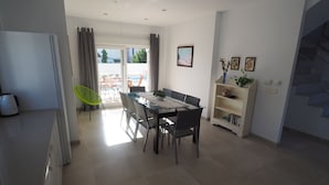 Un comedor con una mesa, sillas y una estantería. Hay una ventana con cortinas, una puerta que da al exterior y una escalera.