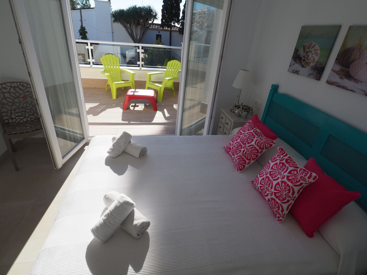 A bedroom with a bed, pillows, a nightstand, and a view of the balcony with chairs.