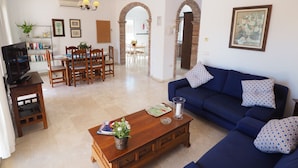 A living room with a blue sofa, wooden coffee table, and a television on a stand. There is a dining area with a table and chairs, and a kitchen in the background. - Nerja Paradise Rentals - Villa Victoria (Nerja)
