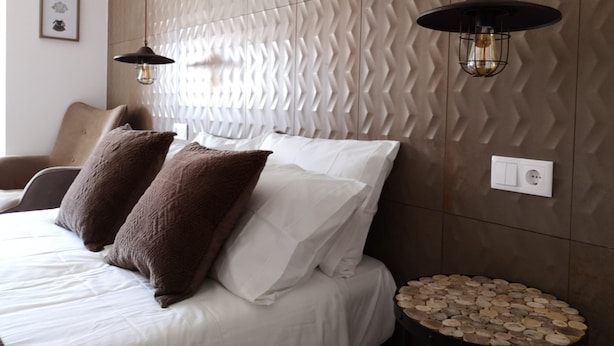A modern bedroom with a textured wall, a bed with white and brown bedding, two pendant lights, and a small round table with wooden objects. - Guilhim Guest House (Nazaré)