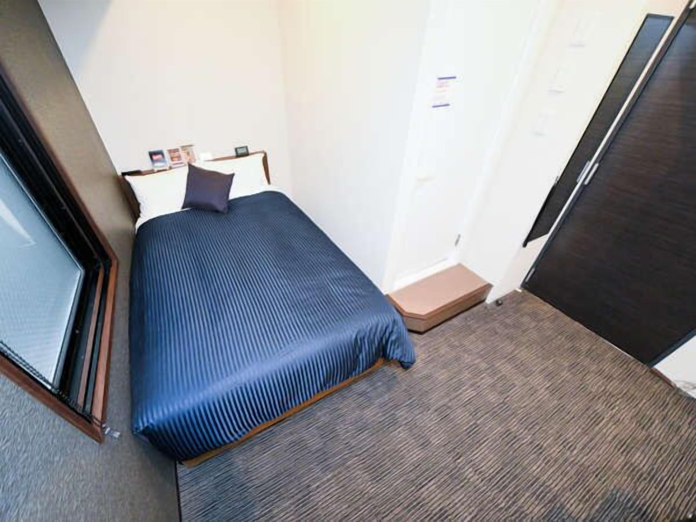 Une petite chambre d’hôtel avec un lit simple, une fenêtre, une porte et un sol recouvert de moquette.