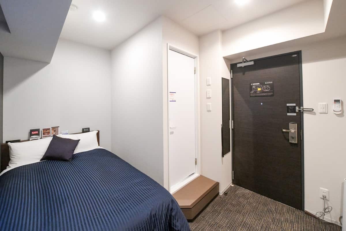 A hotel room with a bed, a door, a small step leading to the door, and a wall-mounted control panel.