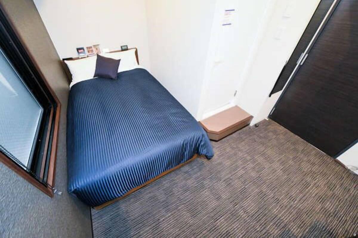 A small hotel room with a single bed, a window, a door, and a carpeted floor.