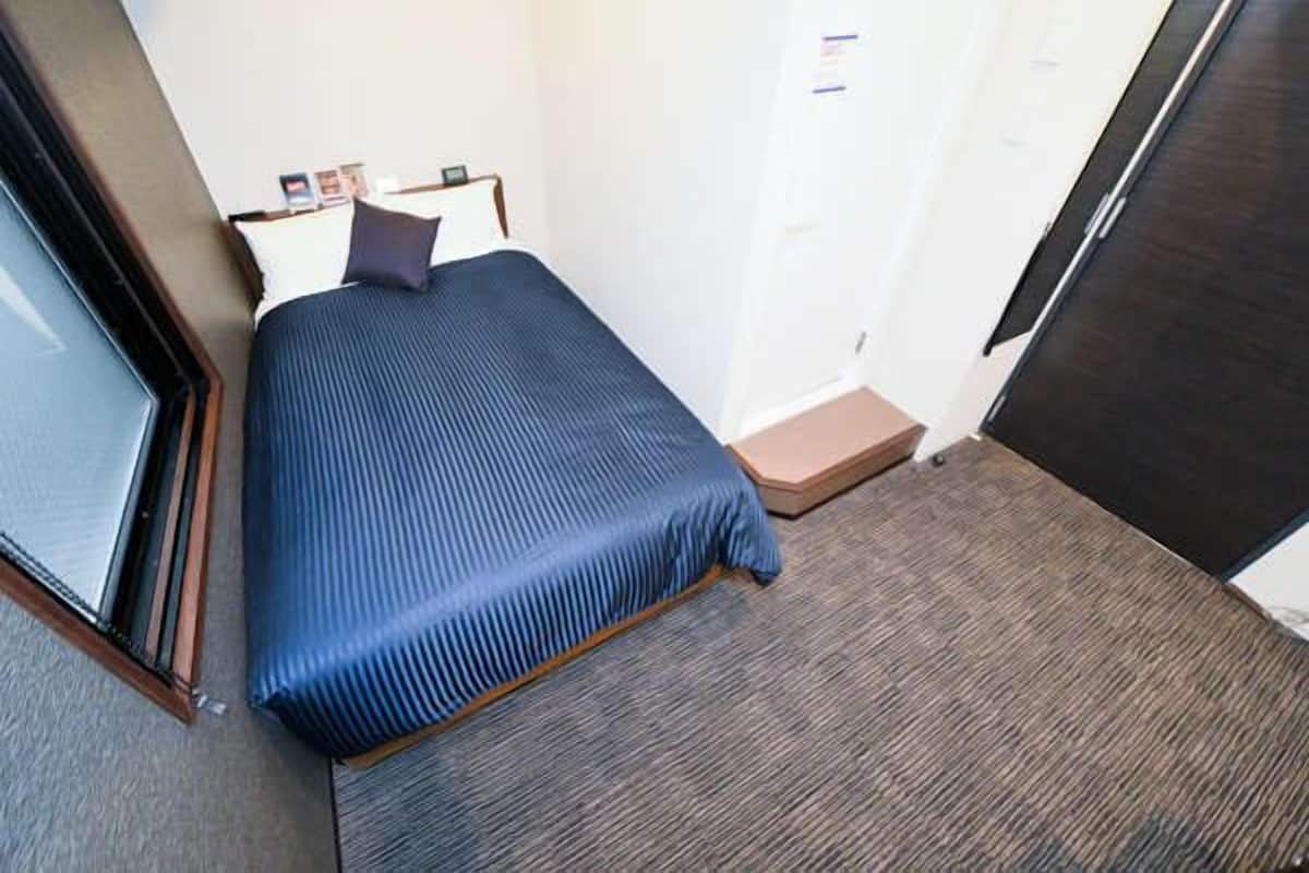 Una habitación de hotel pequeña con una cama individual, una ventana, una puerta y suelo alfombrado.