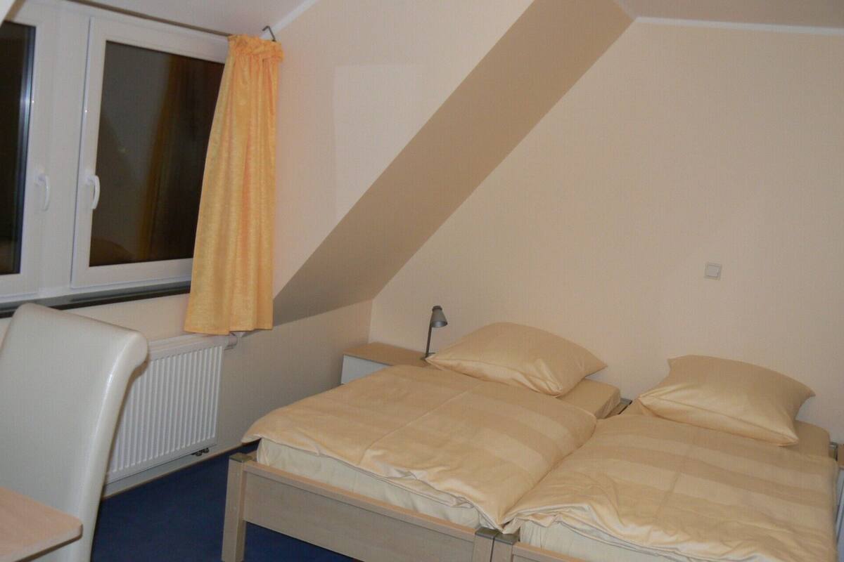 A small, simple room with a bed, a chair, and a window with curtains.