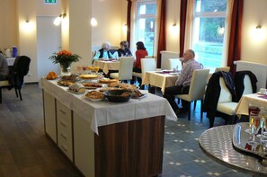 A buffet setup in a dining area with multiple tables and chairs, a central serving station with various dishes, and a window view. - Hotel Appart (Osnabrueck)