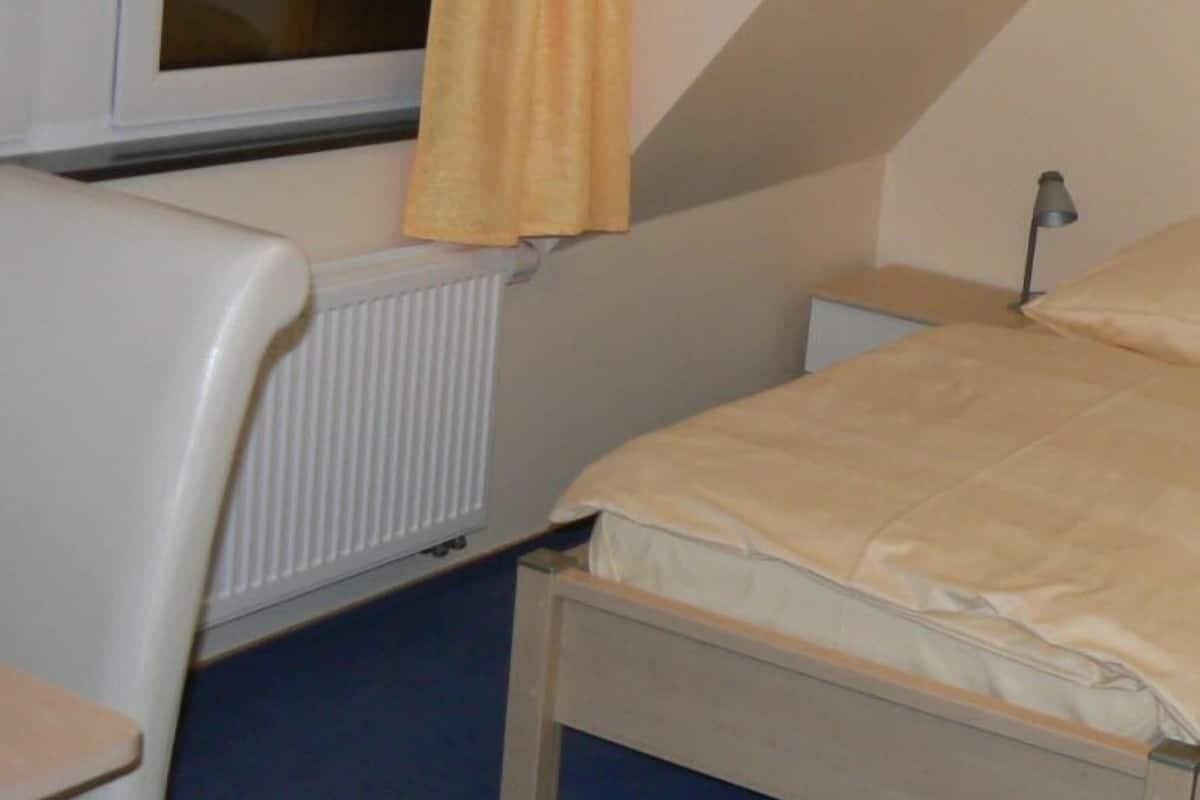 A small, single-bed room with a window, a radiator, and a bedside table.