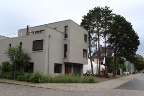 A modern building with a flat roof, multiple windows, and a paved area with greenery in front.