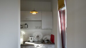 Une cuisine compacte avec des meubles blancs, un seul luminaire suspendu, une fenêtre avec des stores et un petit plan de travail sur lequel se trouvent une bouilloire et une plante.