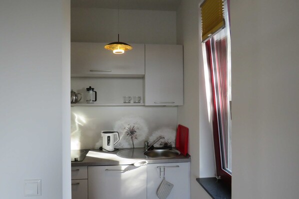 A compact kitchen with white cabinets, a single hanging pendant light, a window with blinds, and a small countertop with a kettle and a plant.