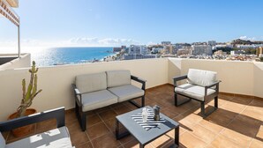 A balcony with a sofa, chair, and table, overlooking a coastal cityscape with buildings and the sea.
