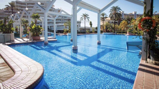A swimming pool with a tiled deck, surrounded by white structures and palm trees.