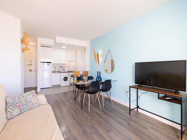A modern living room with a sofa, a dining table with chairs, a TV on a stand, and a kitchen in the background.