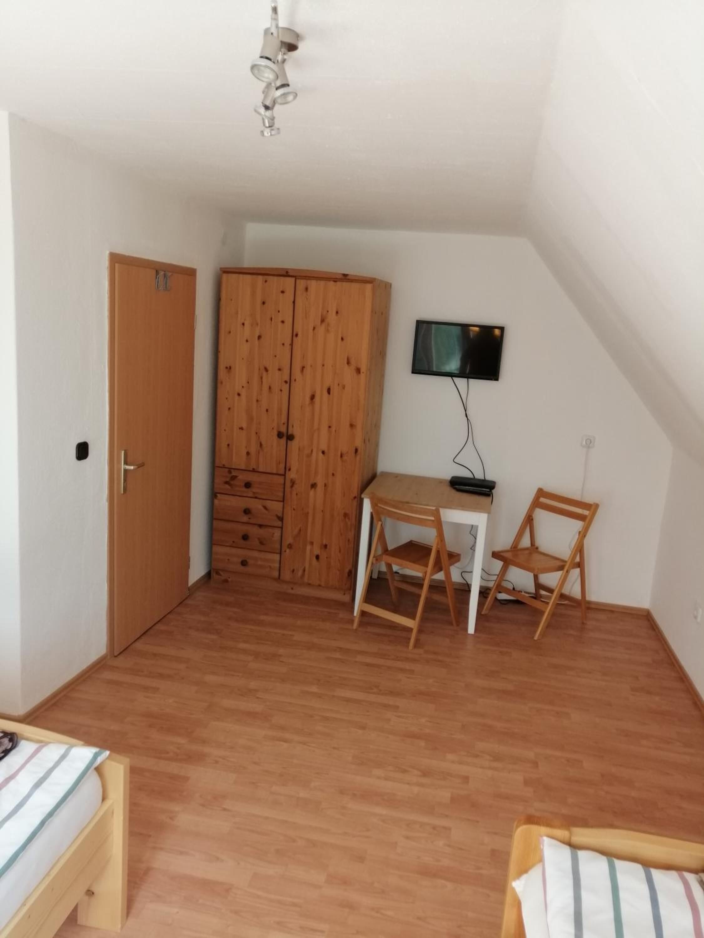 A small, tidy room with a wooden wardrobe, a desk with a chair, a television, and a bed.
