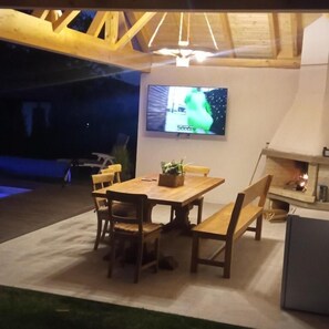 An outdoor dining area with a wooden table and chairs, a flat-screen TV mounted on the wall, and a fireplace.