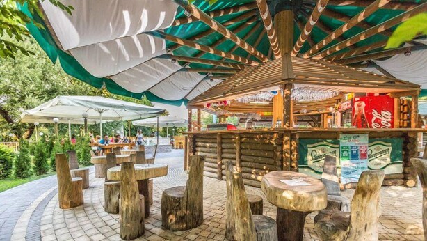 A rustic outdoor seating area with wooden tables and benches, surrounded by a wooden structure with a Coca-Cola advertisement. - Kamp Slapic Mobile Homes (Duga Resa)
