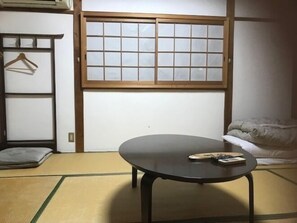 A tatami mat room with a low table, a futon bed, a sliding shoji screen, and a wooden shelf with a hanging item. - Yamashiroya (Nara)