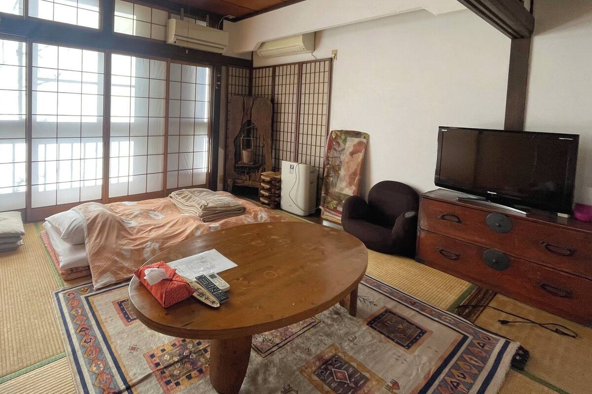 A traditional Japanese room with tatami flooring, a tatami mat with a red cloth, a wooden table, a television, and a sliding door with a grid pattern.
