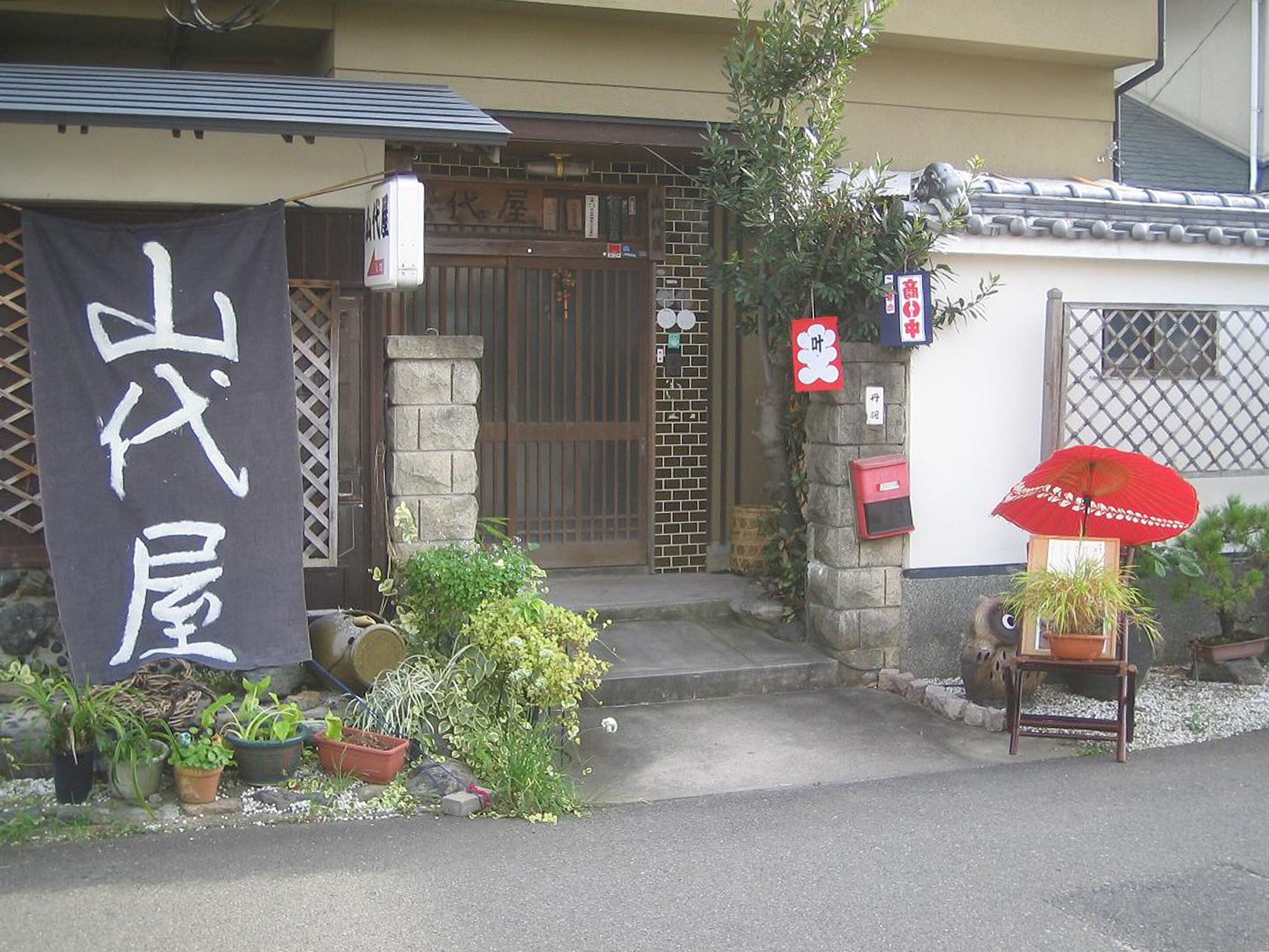 A traditional Japanese house with a wooden gate, a black banner with white characters, and potted plants in front.