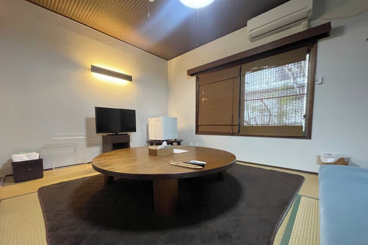 A round table with a remote control and tissues, a flat-screen TV, and a small refrigerator in a room with tatami mats, a window with bamboo blinds, and an air conditioning unit.