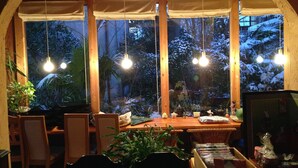 A cozy dining area with a wooden table and chairs, surrounded by large windows with white curtains, overlooking a snowy garden. - Yamashiroya (Nara)