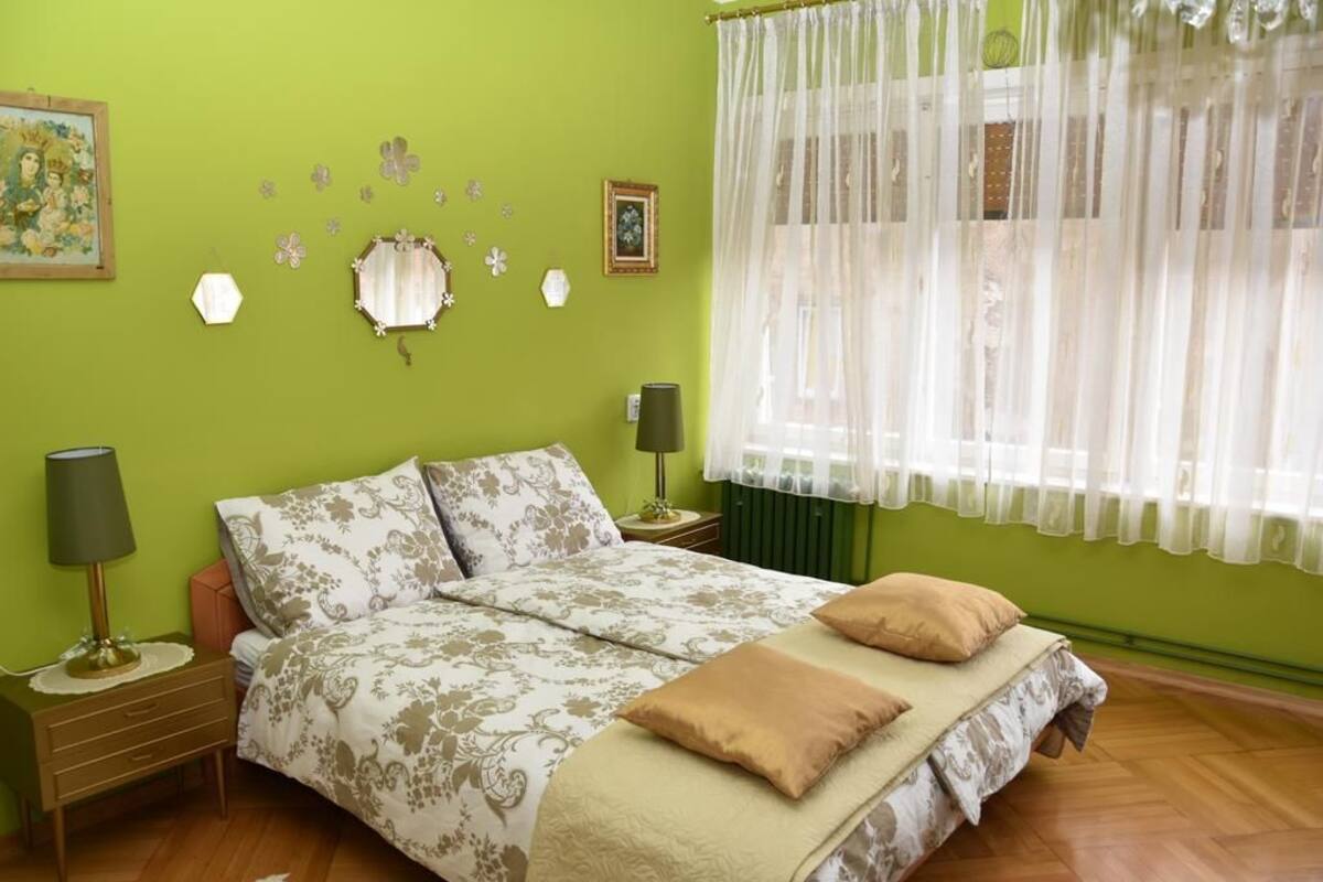Ein Schlafzimmer mit einem Bett, zwei Nachttischen, einem Fenster mit durchsichtigen Vorhängen und Wanddekorationen.