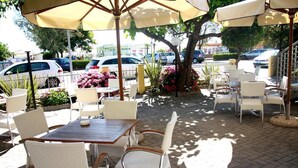 Een terras buiten met witte wicker stoelen en tafels, beschaduwd door grote parasols, omgeven door potplanten en bloemen.
