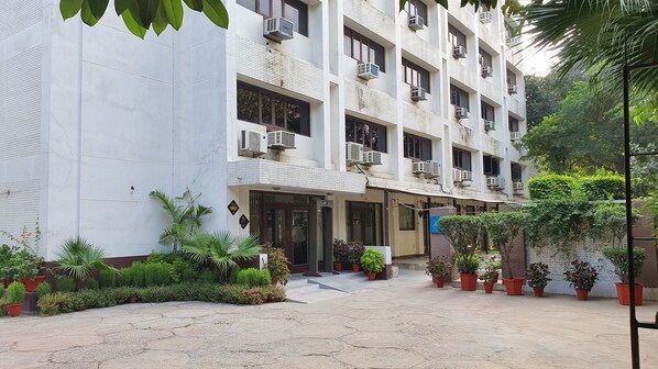 Un edificio de varios pisos con unidades de aire acondicionado en el exterior, una entrada con portones y plantas en macetas alrededor del acceso.