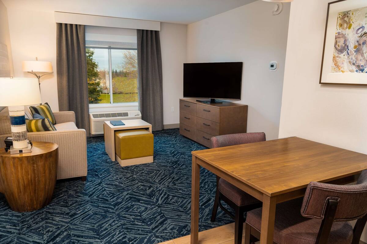 Een hotelkamer met een bank, een houten bijzettafeltje, een televisie op een kast, een eettafel met stoelen en een groot raam met gordijnen.