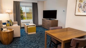 A hotel room with a sofa, a wooden side table, a television on a cabinet, a dining table with chairs, and a large window with curtains.