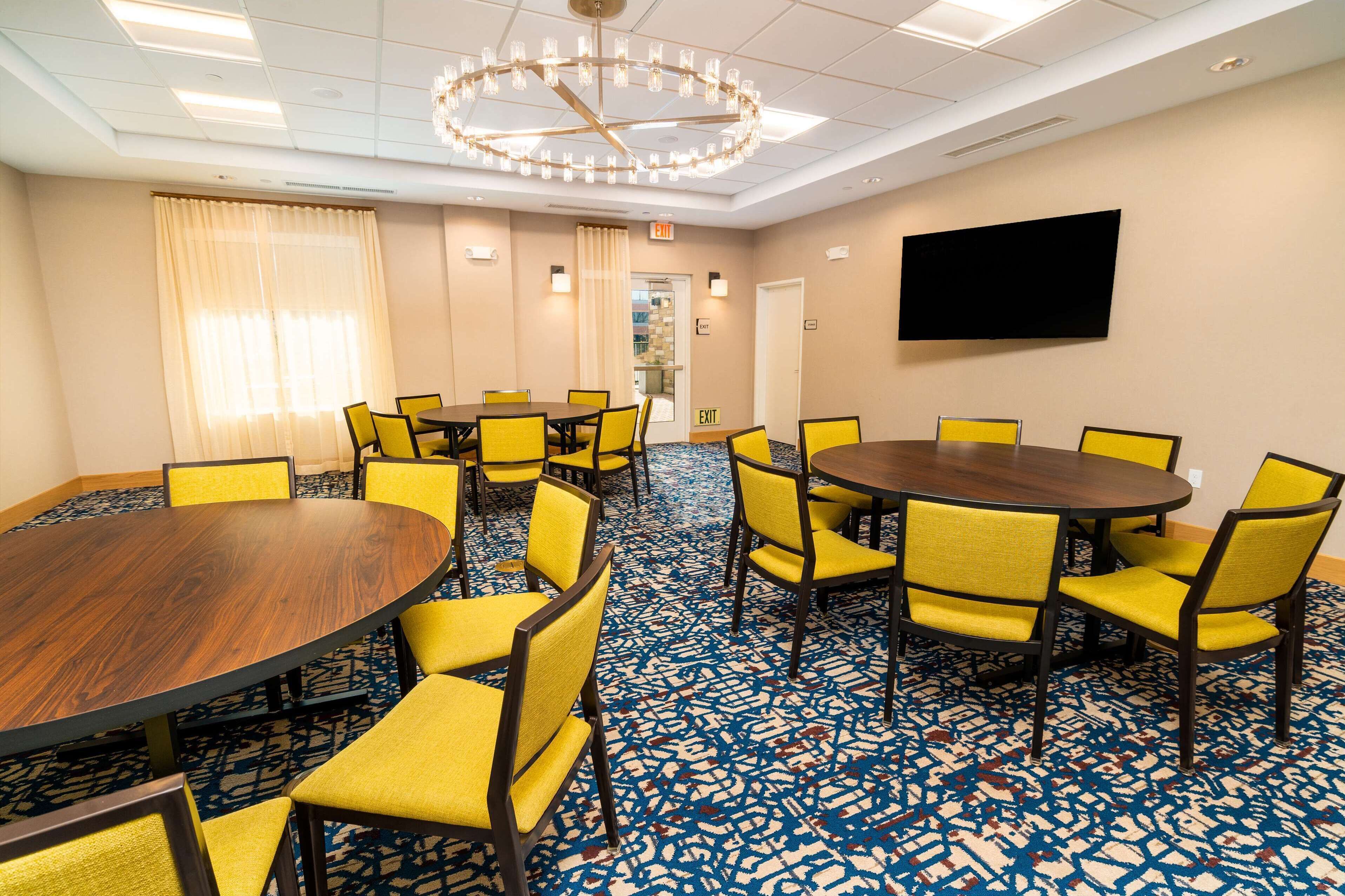A conference room with a large round table, yellow chairs, a patterned carpet, a chandelier, and a flat-screen TV on the wall.