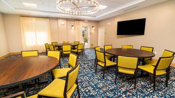 A conference room with a large round table, yellow chairs, a patterned carpet, a chandelier, and a flat-screen TV on the wall.