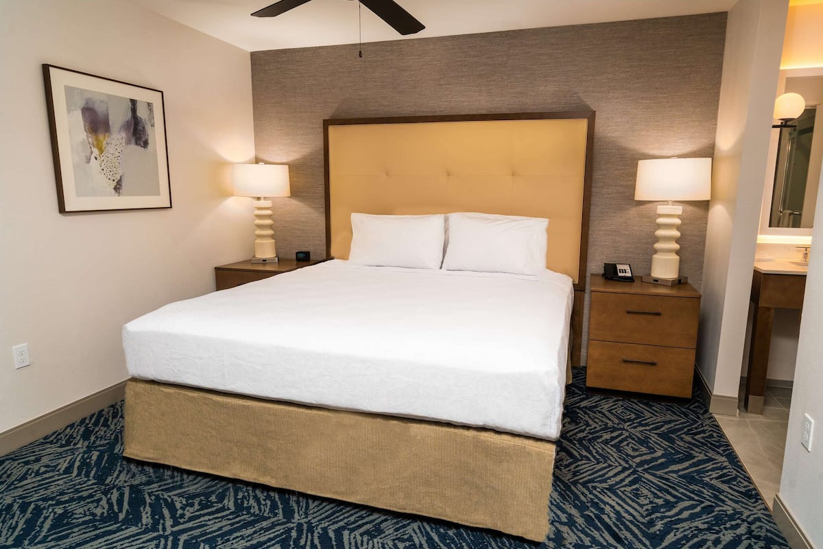 Een hotelkamer met een groot bed, twee nachtkastjes met lampen, een houten hoofdbord, een lijst met een abstract schilderij en een kamerbreed tapijt met dessin.