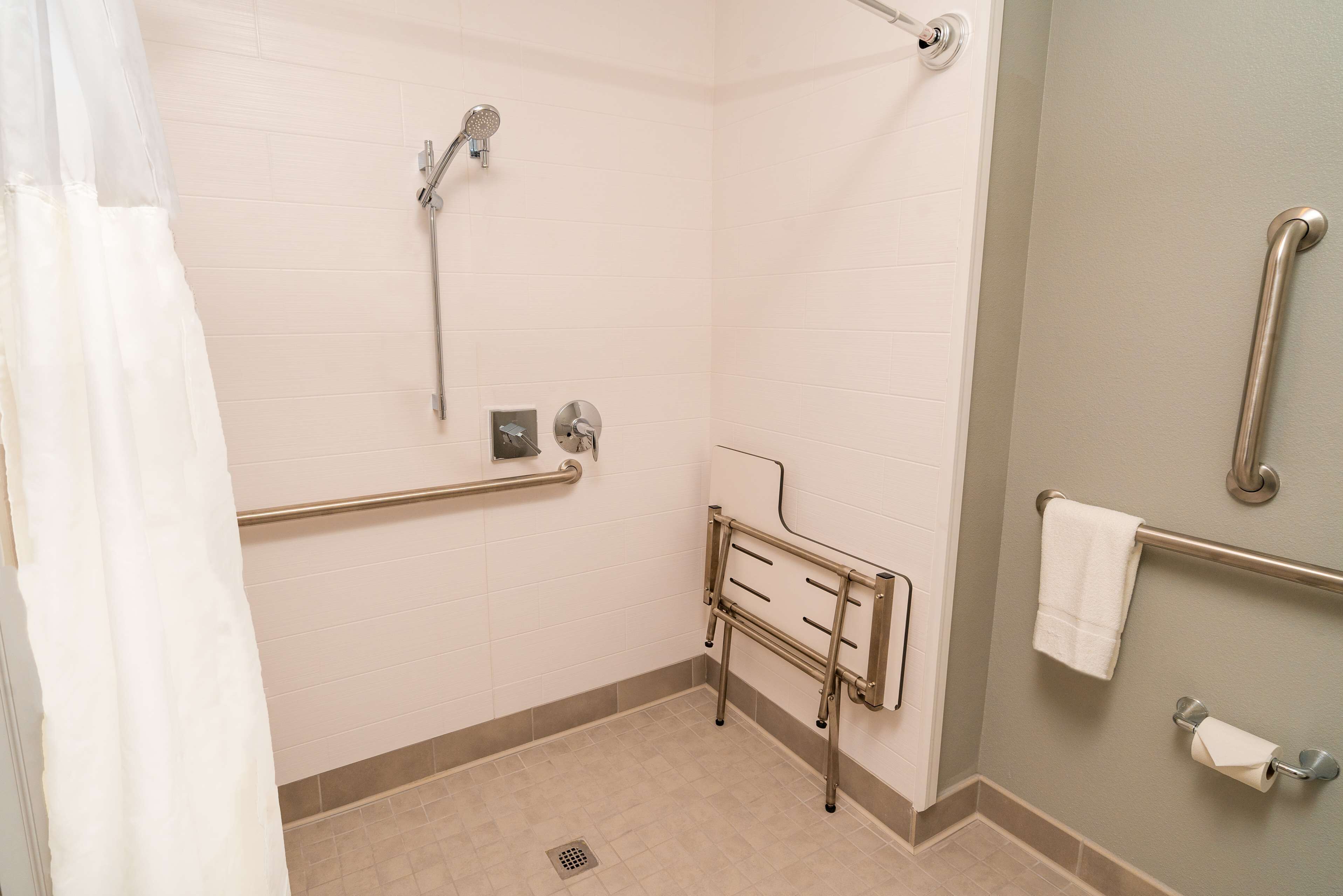 Un baño con ducha, barras de apoyo, secamanos y perchero para toallas.