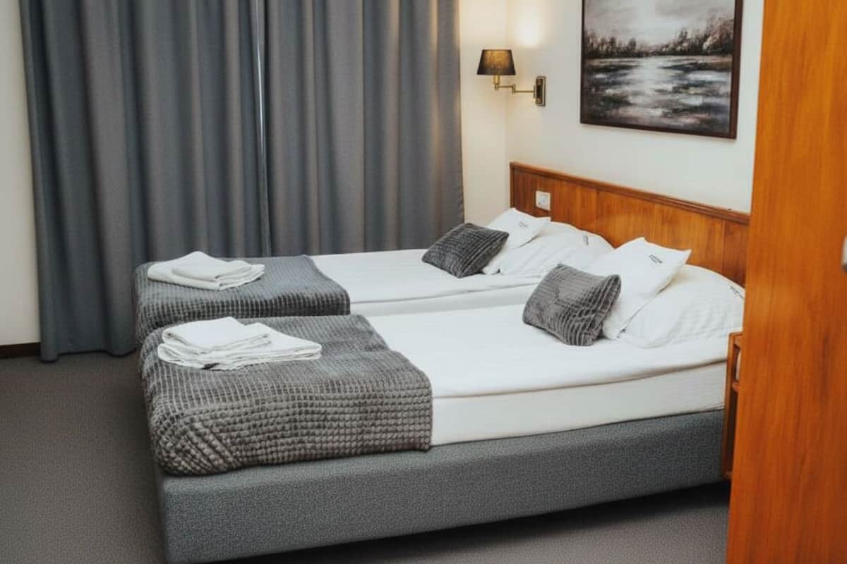 Hotellihuone, jossa on kaksi sänkyä, harmaat vuodevaatteet, puinen pääty ja seinällä taulu sekä lamppu.