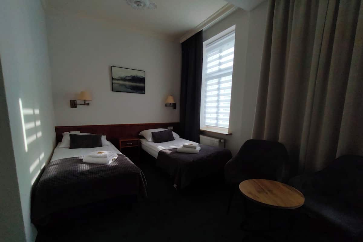 Hotellihuone, jossa on kaksi sänkyä, tuoli, pieni pöytä ja ikkuna, jossa on sälekaihtimet.