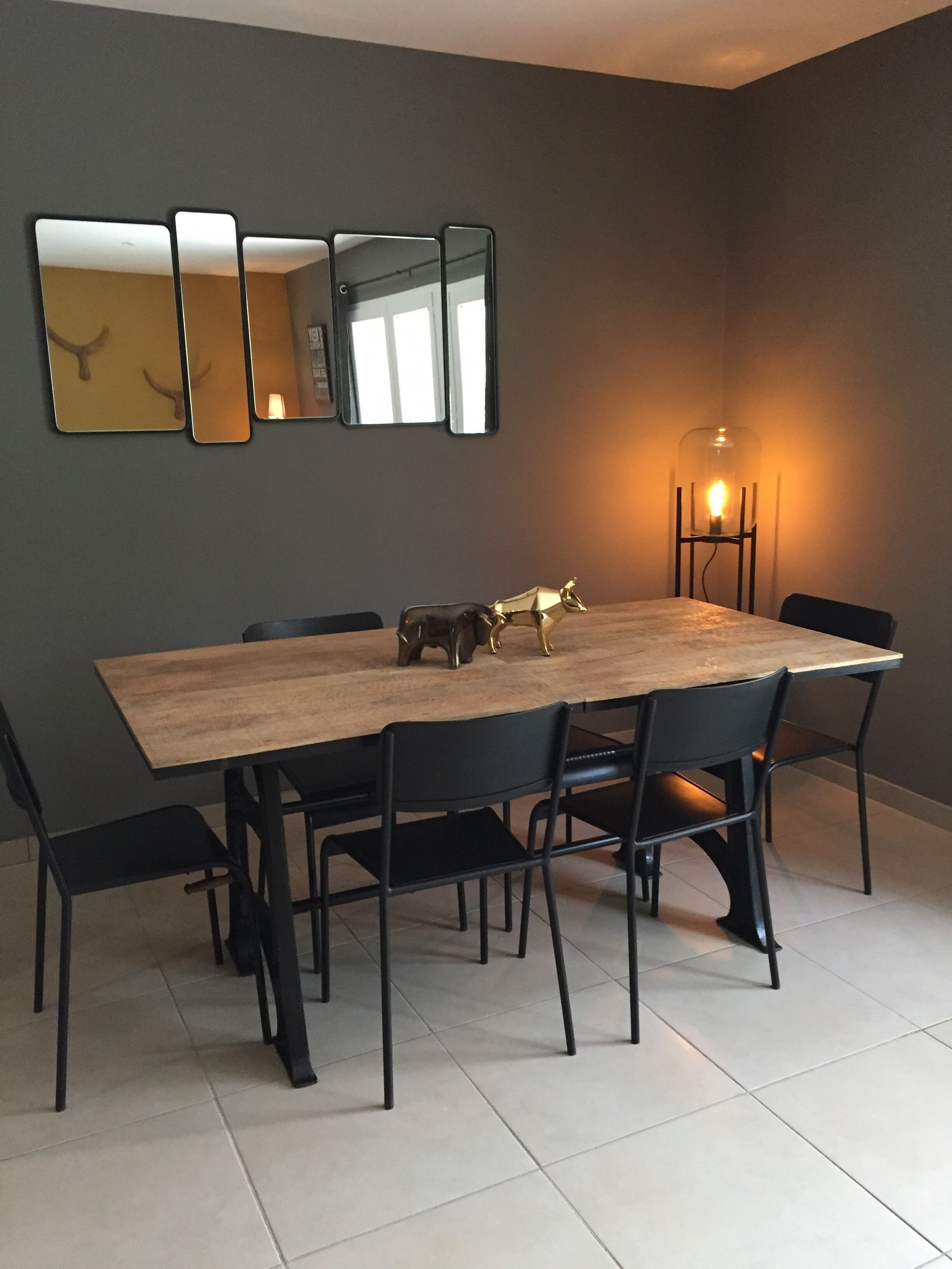 Un coin repas avec une table rectangulaire en bois, entourée de chaises noires. Quatre miroirs sont accrochés au mur au-dessus de la table. Une seule lampe de sol, dotée d'un abat-jour en verre, est visible sur le côté droit.