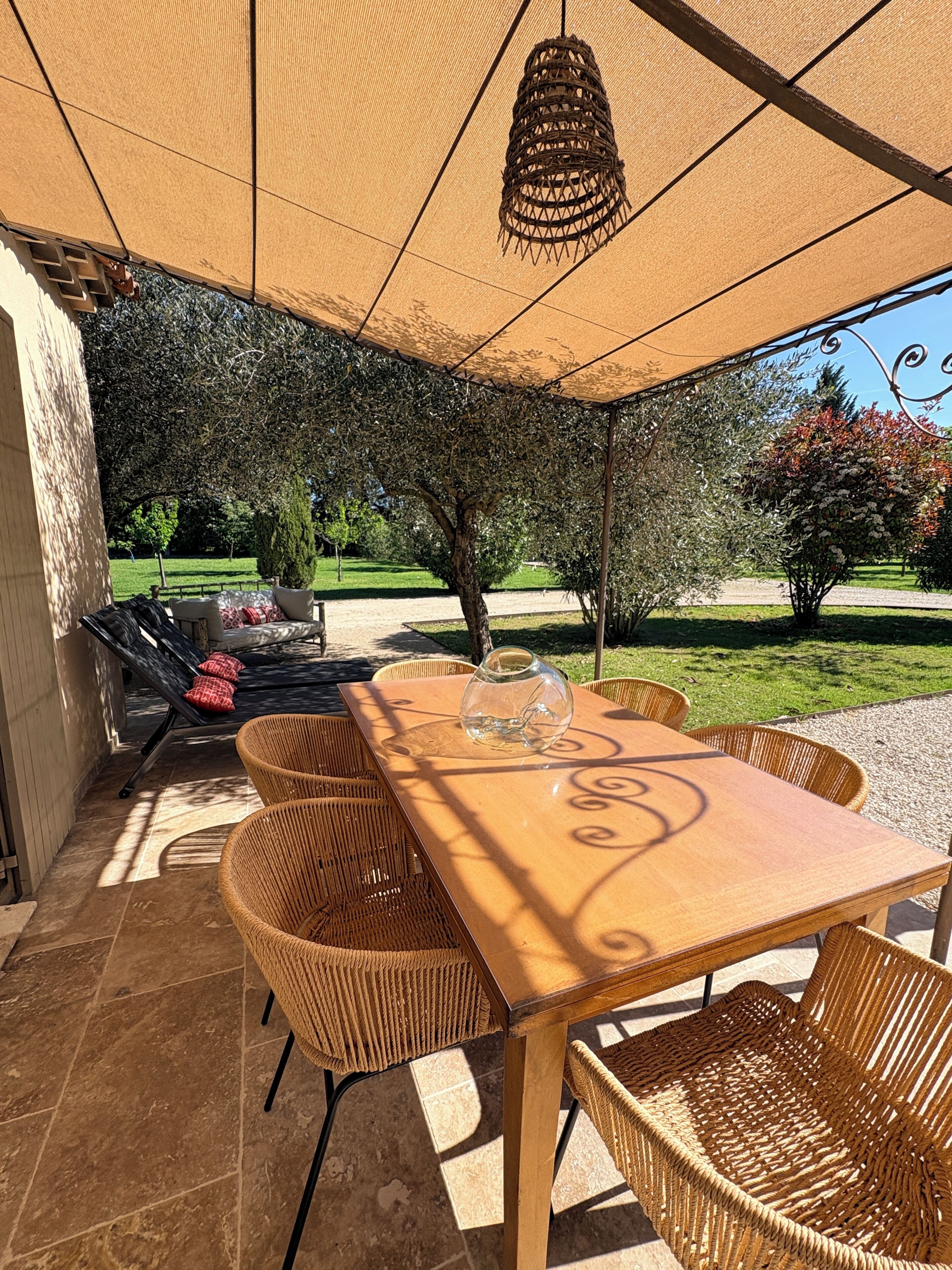 Une terrasse extérieure avec une table et des chaises, un luminaire suspendu et une vue sur un jardin avec des arbres et une pelouse.