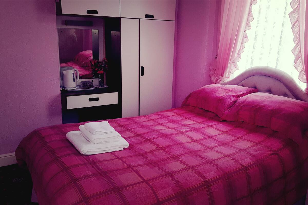 Une chambre avec une couvre-lit rose, une table de chevet blanche, un miroir et une armoire.
