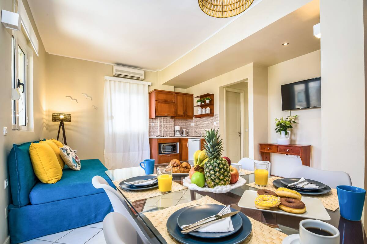 A modern living room with a blue sofa, dining table set with plates, cutlery, and a fruit bowl.