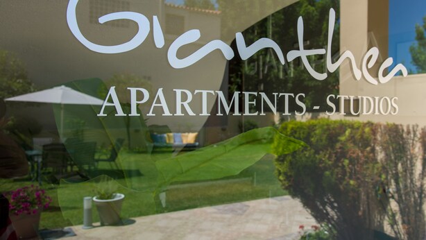A window with the text "Gianthe Apartments - Studios" and a reflection of a building and greenery. - Gianthea Studios and Apt (Chania)