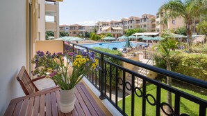 A balcony with a wooden table and a vase of flowers, overlooking a swimming pool area with umbrellas and palm trees. - Gianthea Studios and Apt (Chania)