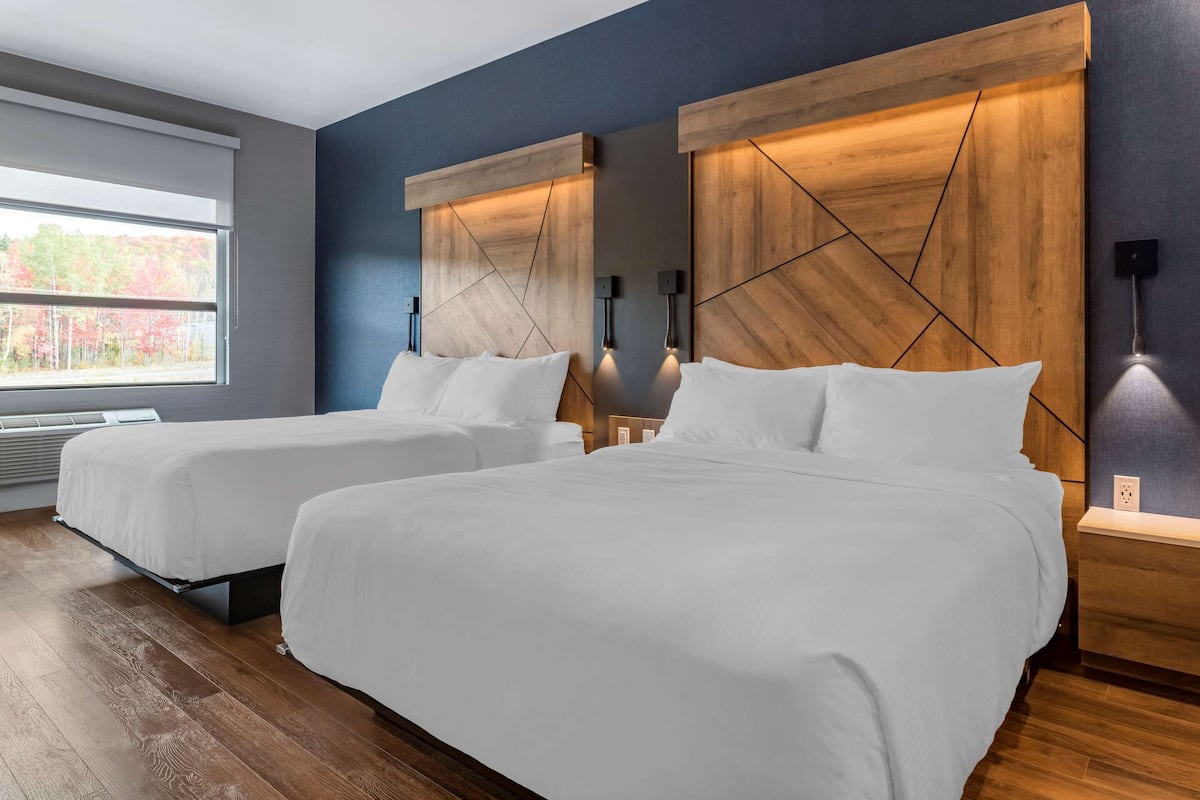 A hotel room with two beds, wooden headboards, bedside lamps, and a window with a view of trees.
