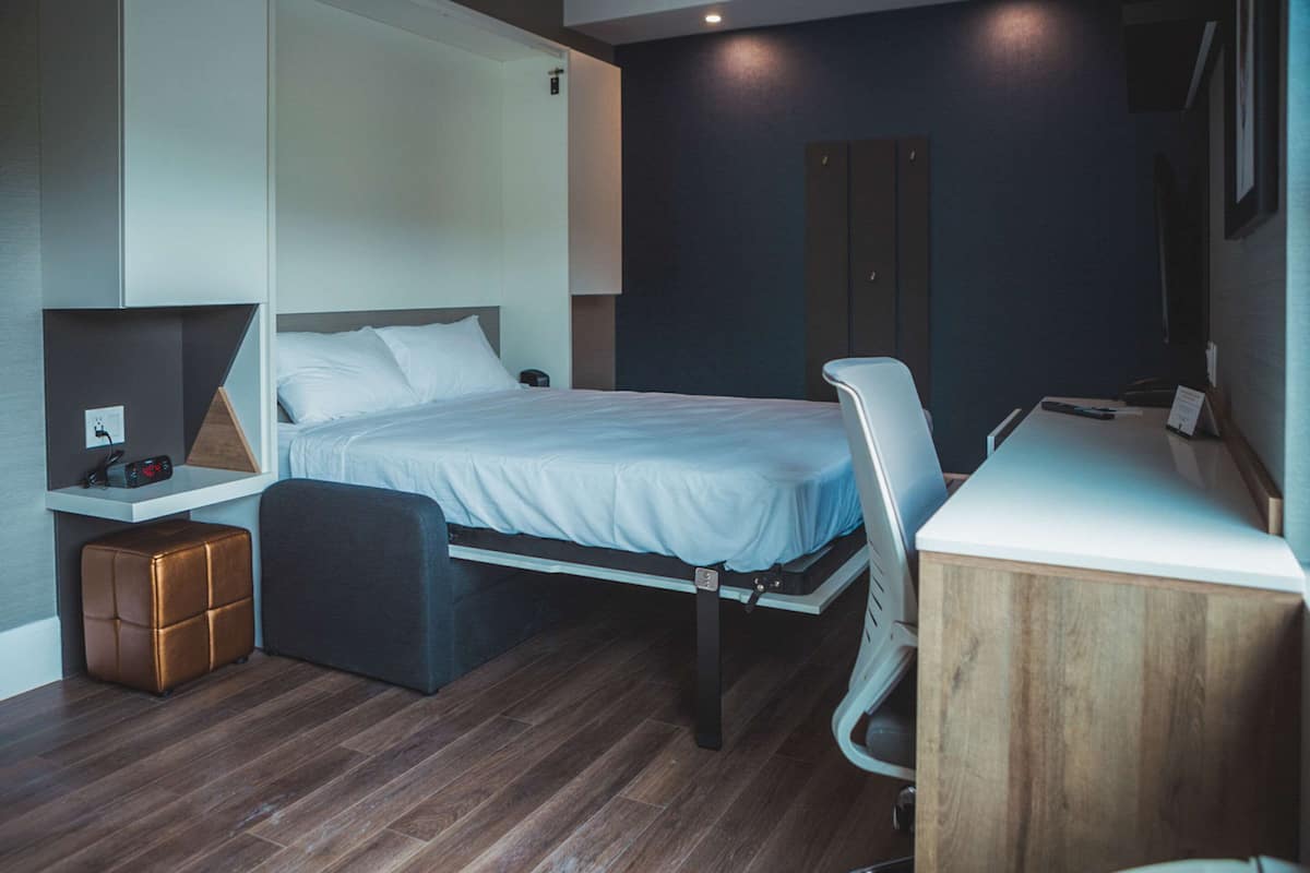 A modern hotel room with a bed, a desk, a chair, and a wardrobe.