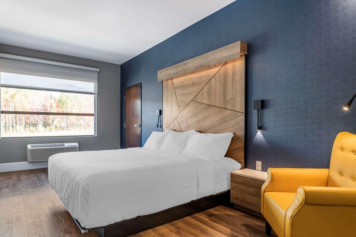 Un dormitorio moderno con una cama grande, cabecera de madera, un sillón amarillo y una ventana con vistas a árboles.